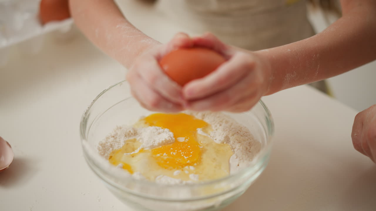 hand view of person breaking brown egg into bowl filled with flour and cracked yolk, preparing ingredients for baking on white surface, showcasing hands-on kitchen
