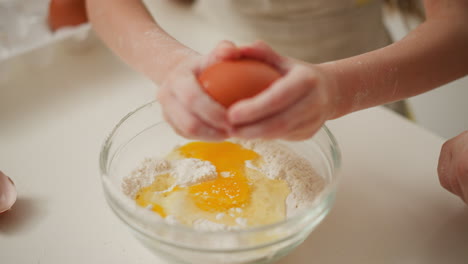 hand view of person breaking brown egg into bowl filled with flour and cracked yolk, preparing ingredients for baking on white surface, showcasing hands-on kitchen