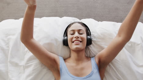mujer biracial feliz acostada en la cama usando auriculares y escuchando música, cámara lenta