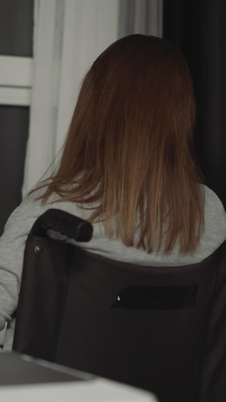 체어에서 마비 된 여성은 부 창문으로 이동합니다. 젊은 주부는 주말 저녁에 평온하게 거리를 바라보며 집에서 휴식을 취합니다.