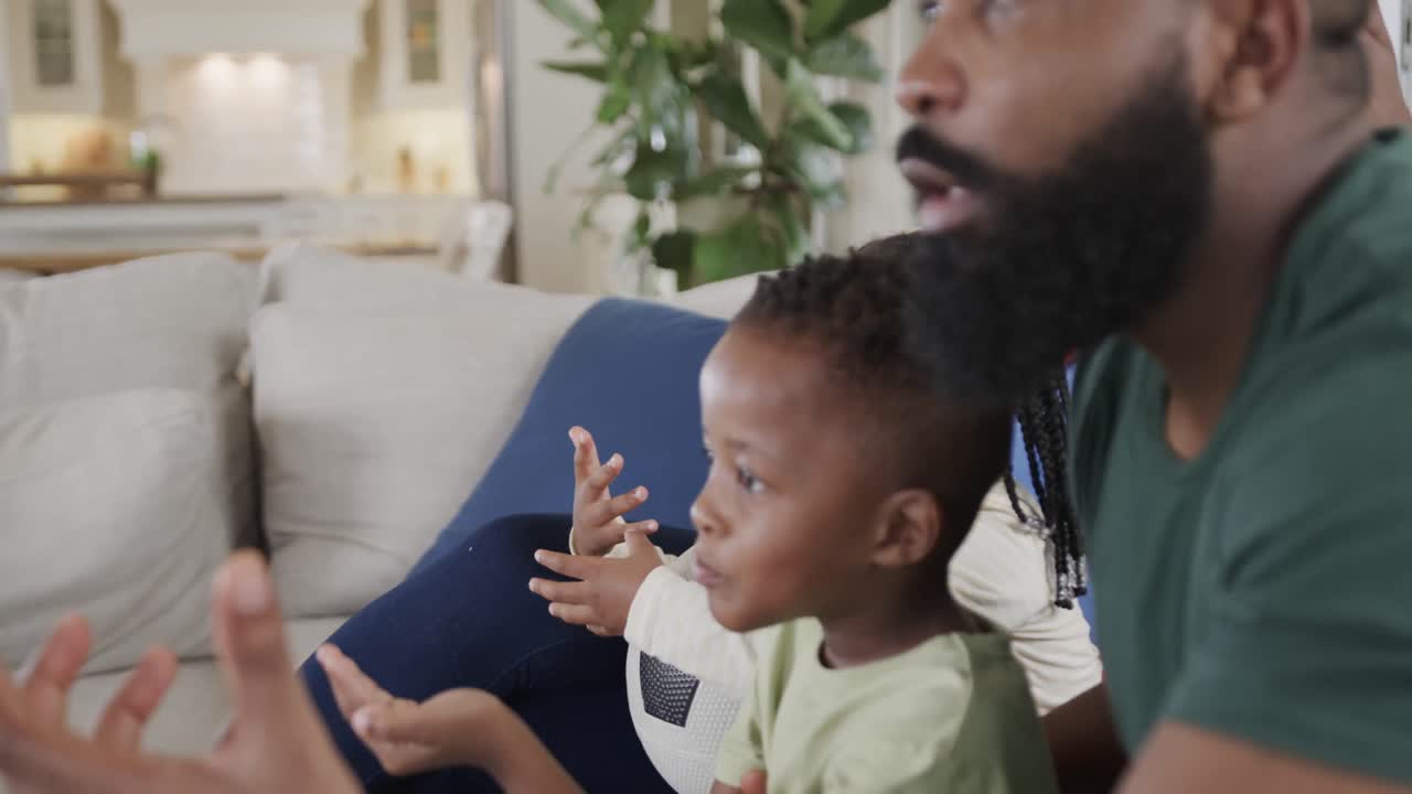 Excited african american parents with son and daughter watching sport on tv at home, slow motion