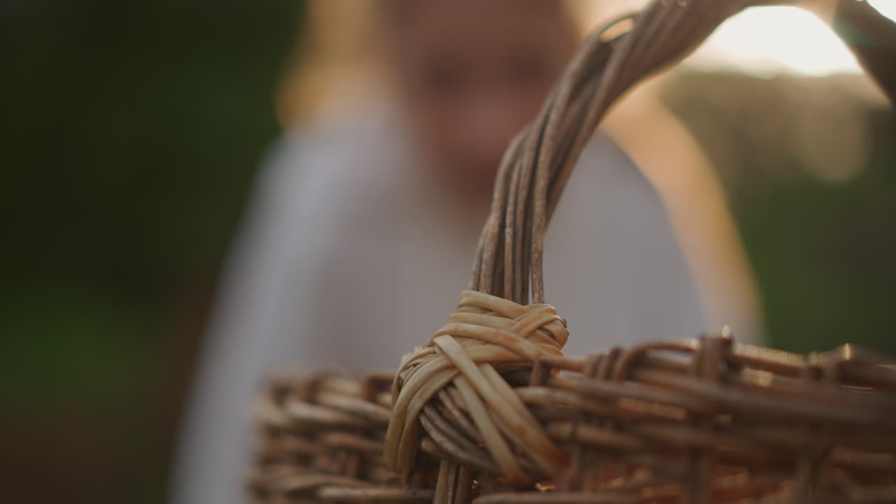 Manico del cesto in primo piano con figura sfocata, bambino che si avvicina al giardino e allunga la mano verso il manico, retroilluminazione soffusa e bokeh creano un bagliore giocoso, dettagli di nodi e spago enfatizzati, picnic familiare spontaneo.