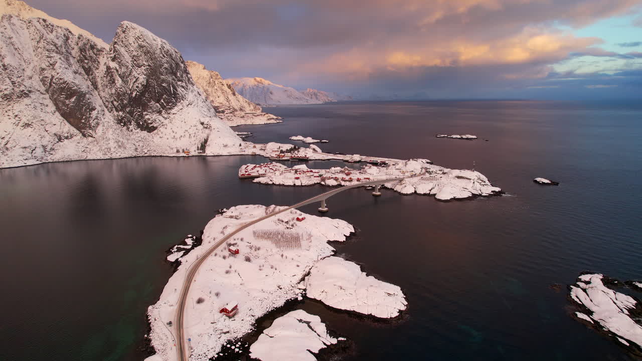 Cinematic 4K drone shot of Hamnoy village at sunset. Iconic red rorbuer cabins against snowy mountain and reine fjord in Lofoten, Norway. Winding E10 road and bridge conecting small islands
