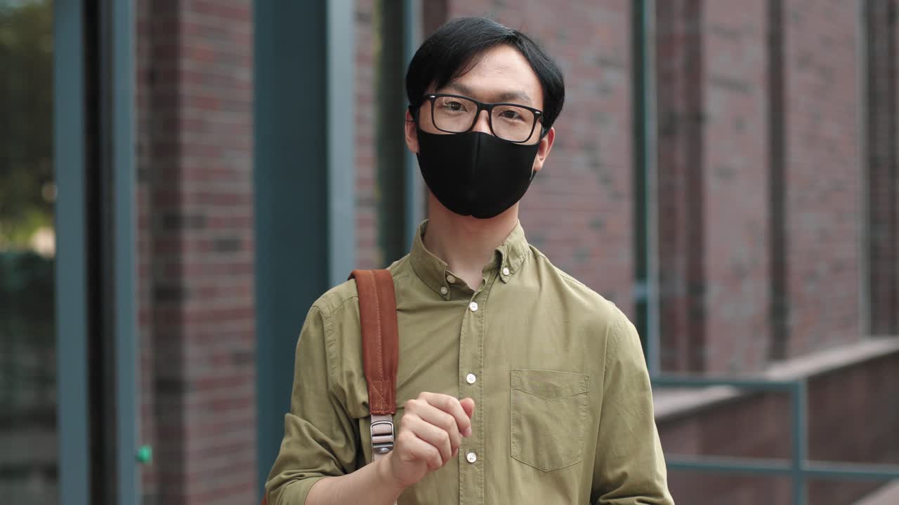대학 근처에서 카메라를 보면서 안경을 쓰고 스마트폰을 들고 있는 안면 마스크를 쓴 아시아 학생을 카메라로 확대