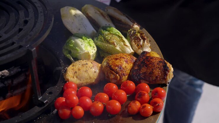 Primo piano di una piastra braciere con verdure e carne in cottura, un uomo si avvicina per aggiungere un pizzico di condimento