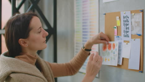 young woman colorist designer picks up colors standing near the wall. looking at the wall of sketches and flowers. choose the right color.