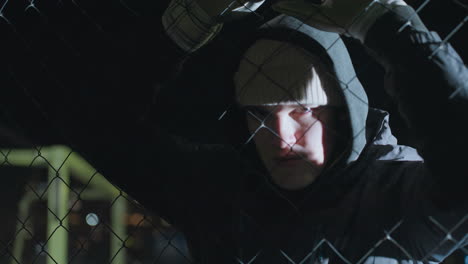 young man resting on mesh-like fence under bright light, tired expression illuminated by reflections, wearing gloves and hooded jacket, with blurred bokeh lights