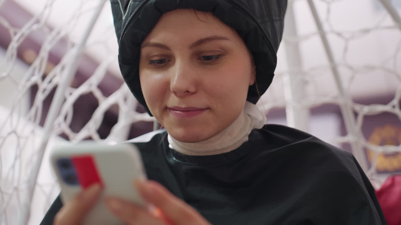 미용실 의자에 앉아 시술 랩을 두른 채 휴대폰을 보며 휴식을 취하는 백인 고객, 케이프를 두르고 망사 후드를 덮어쓴 모습, 시술 중 차분한 휴식 시간, 은은한 조명과 현대적인 인테리어, 편안한 분위기