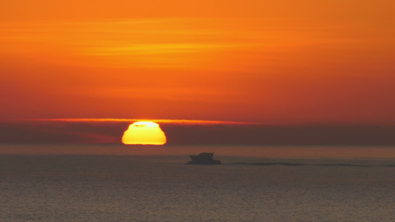 Dramatic slow motion sunrise over ocean. Small motorboat silhouette, orange sun on horizon.