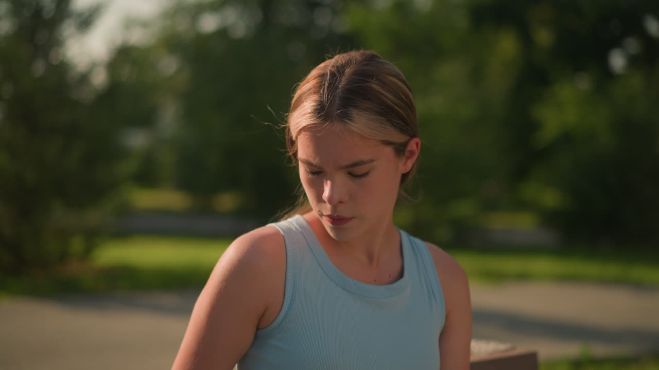 은 날에 야외에 앉아있는 젊은 백인 여성은 푸른 크 을 입고 있으며, 그녀의 표정은 집중적이고 평온하며, 흐릿한 배경은 녹색 나무와 잎자루를 특징으로합니다.