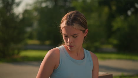 joven dama blanca mirando pensativa hacia abajo mientras está sentada al aire libre en un día soleado, lleva una camiseta azul, su expresión enfocada y tranquila, el fondo borroso presenta árboles verdes y follaje