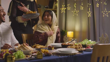 familia musulmana sentada alrededor de una mesa en casa con una mujer sirviendo biryani en la comida para celebrar el eid 1