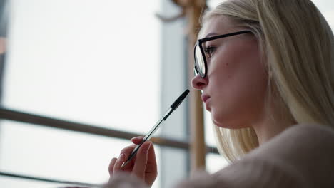 primer plano de una mujer blanca reflexiva golpeando suavemente la pluma en sus labios mientras está profundamente pensada, lleva gafas con un fondo borroso, transmitiendo un momento reflexivo e introspectivo