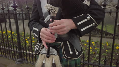 close-up of a male scottish bagpipers hands as he plays the bagpipes next to a black, ornate metal fence with princes street gardens in the background, edinburgh, scotland