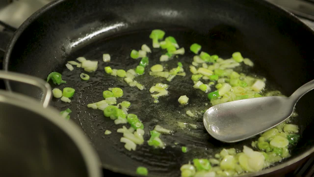 Close-up of chopped onions and green peppers being saut&eacute;ed in a hot pan with oil, stirred by a metal spatula, kitchen setting