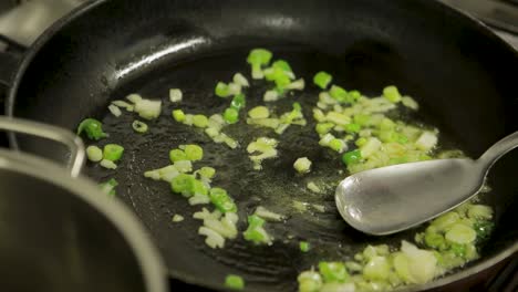 close-up of chopped onions and green peppers being sautéed in a hot pan with oil, stirred by a metal spatula, kitchen setting