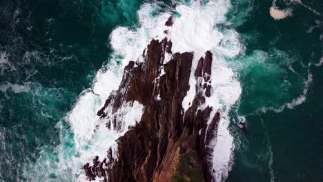 blue ocean waves crash onto sharp rocks, rising ultra wide drone shot