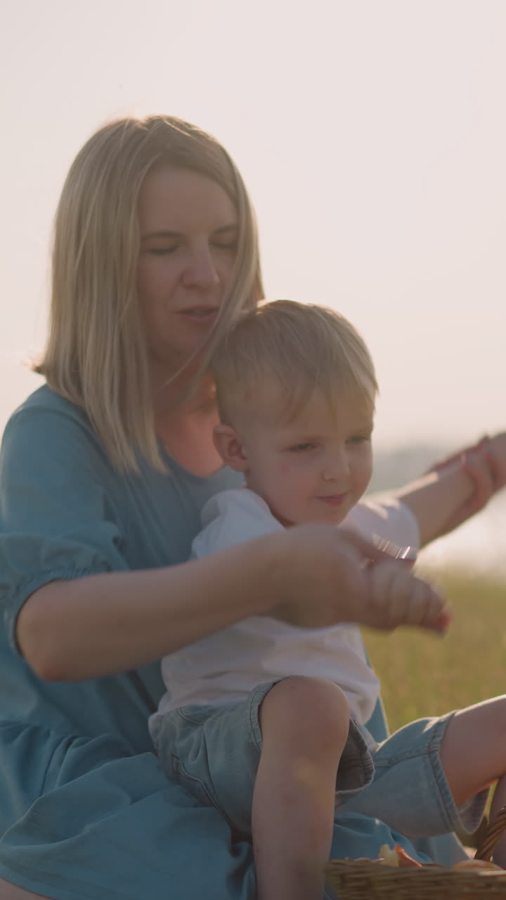 A caring mother in a blue dress with her young son on her lap, gently holding his hands as they sit in the grass. His older brother, holding an apple, in the background near a basket of fruit