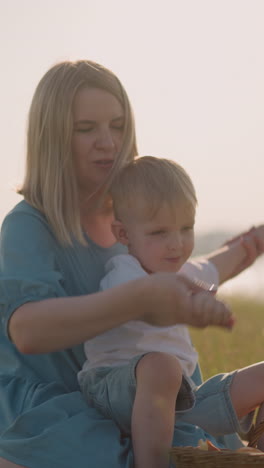 a caring mother in a blue dress with her young son on her lap, gently holding his hands as they sit in the grass. his older brother, holding an apple, in the background near a basket of fruit