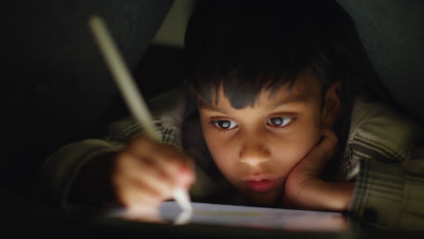 close up of young boy in home made camp made from cushions playing and drawing with digital tablet and stylus at night 3