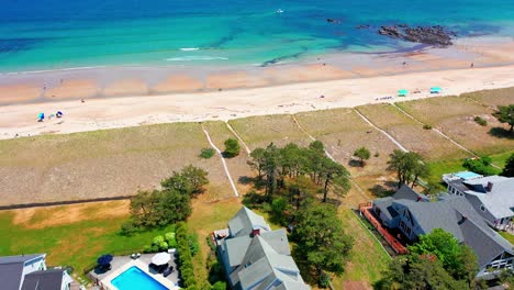 drone shows saco, maine’s beach bustling with tourists. sand full of people sunbathing, swimming, and playing near vacation homes as blue atlantic waves wash over the new england seashore