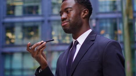 Close-Up-Of-Young-Businessman-Wearing-Suit-Talking-On-Mobile-Phone-Using-Built-In-Microphone-Standing-Outside-Offices-In-The-Financial-District-Of-The-City-Of-London-UK-Shot-In-Real-Time-1