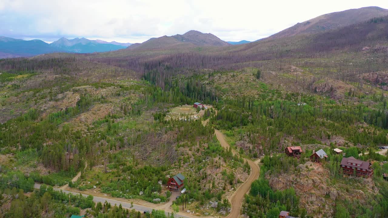 Veduta aerea di una valle di montagna con baite sparse, strade sterrate tortuose, ricrescita forestale, pendii rocciosi e vaste pendii di alberi morti sotto un cielo coperto in un paesaggio selvaggio e remoto