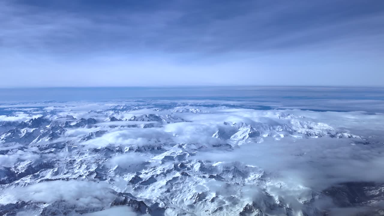 Hyperlapse kuvattuna suihkukoneen ohjaamosta Alppien yli lennettäessä. Vuorenhuiput ovat osittain hajanaisten pilvien peitossa sinisen taivaan alla.