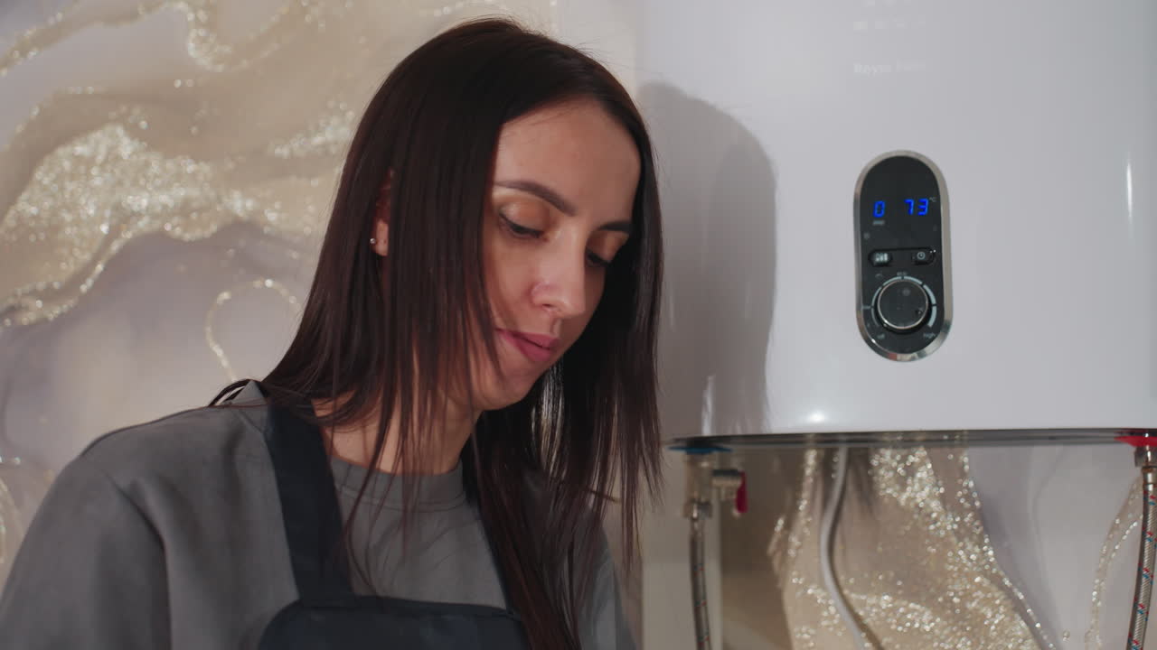 La dueña o gerente caucásica de un salón de belleza comprueba el calentador de agua y las conexiones en el trastero cerca del lavabo, con expresión de preocupación mientras prepara el equipo para el servicio. Se ve una unidad industrial y un panel de control.