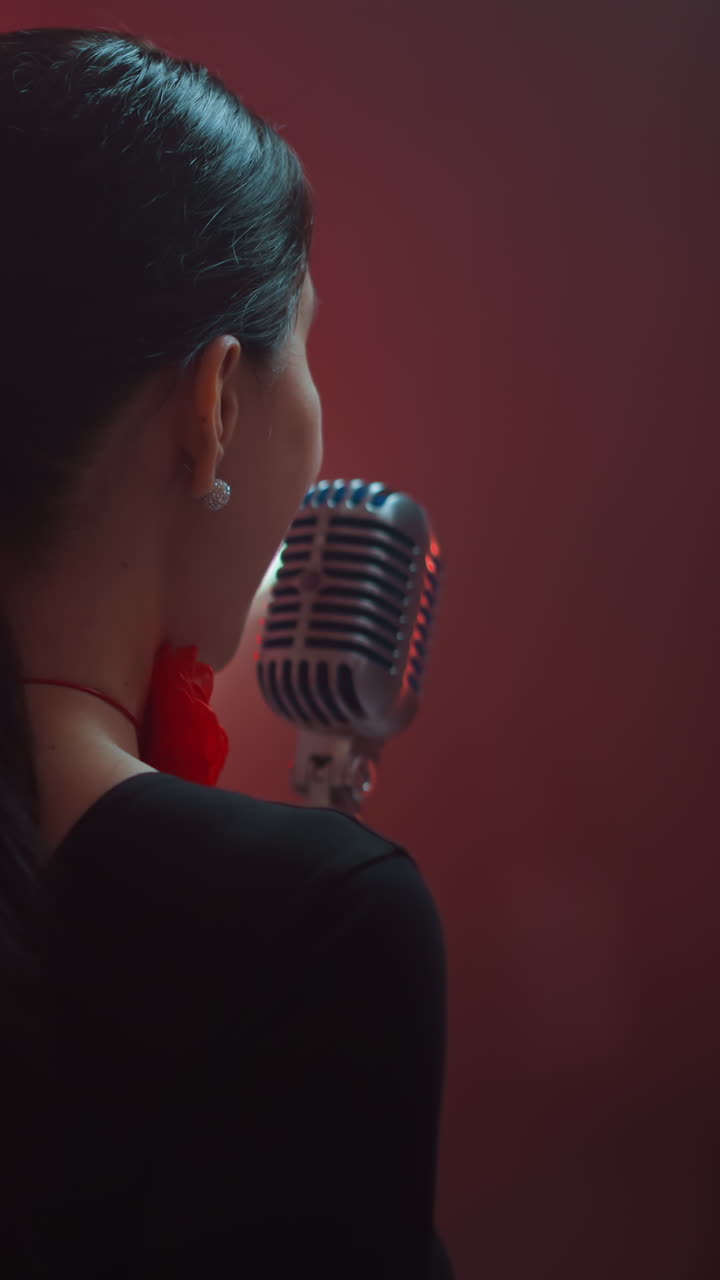 vista trasera de una cantante con una cinta roja en el cuello, cantando en un micrófono vintage con un guitarrista en el fondo rojo borroso