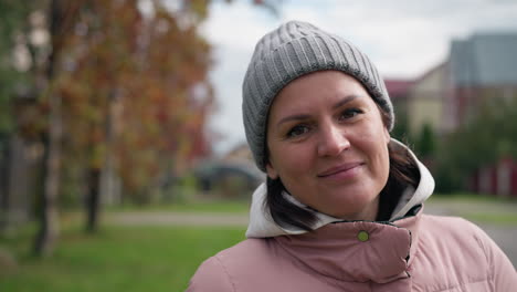 mujer de mediana edad con una cálida sonrisa inclina su cabeza ligeramente mientras está de pie al aire libre, con un fondo borroso de árboles coloridos y un ambiente sereno y tranquilo