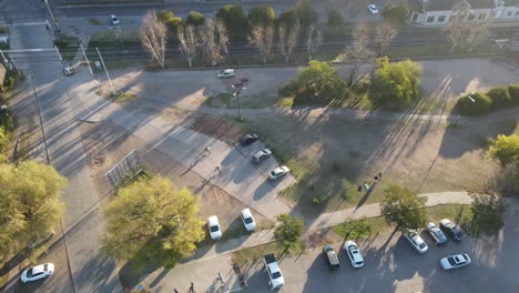 Aerial-view-of-a-couple-of-cyclists-riding-through-a-park,-with-several-cars-parked-around-it-in-the-sunset-light