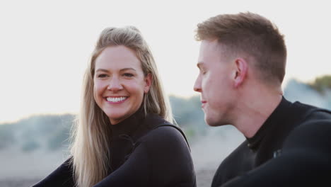 couple wearing wetsuits sitting on beach talking and looking out to sea together