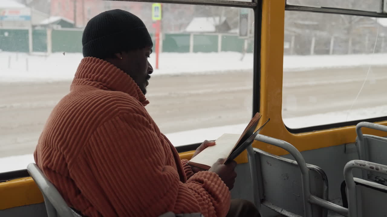 Black man reading on bus, engrossed in paperback during chilly urban commute, wearing orange knit coat and black beanie, snow visible outside window, empty seats and muted street traffic, reflective