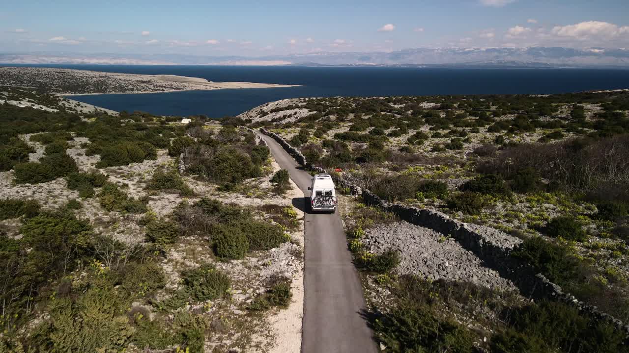 plano recto con un dron detrás de una autocaravana a baja altura mirando el mar en el horizonte cielo azul y verde en las montañas día soleado 4k