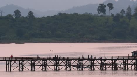 Mon-Bridge-and-a-longboat-followed-speeding-behind-the-structure-while-people-are-walking-on-the-bridge,-silhouetting-as-it-was-getting-dark,-in-slow-motion