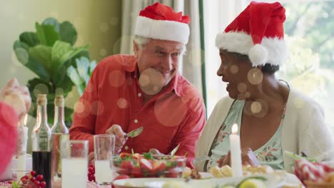 animación de puntos de luz sobre una pareja mayor diversa con sombreros riendo en la mesa de la cena de navidad