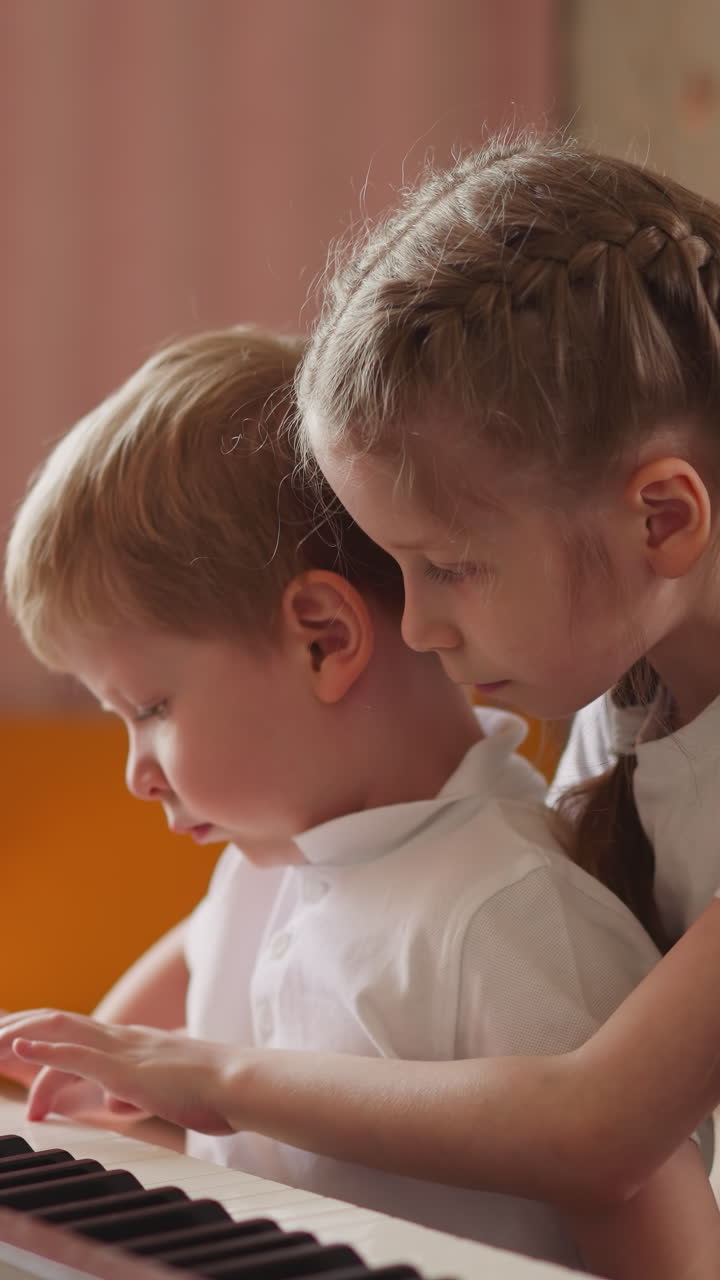 Girl with orthopedic back braces plays piano hugging toddler blond boy in living room. Curious little brother looks at sister hands on instrument keyboard