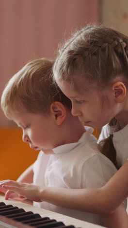girl with orthopedic back braces plays piano hugging toddler blond boy in living room. curious little brother looks at sister hands on instrument keyboard