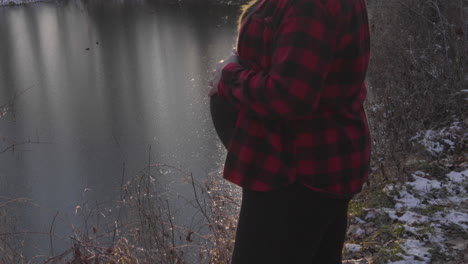 a tilt up shot of a pregnant woman in her third trimester stands holding her baby belly as she gazes across a pond in the woods on a winter day with a dusting of snow on the ground