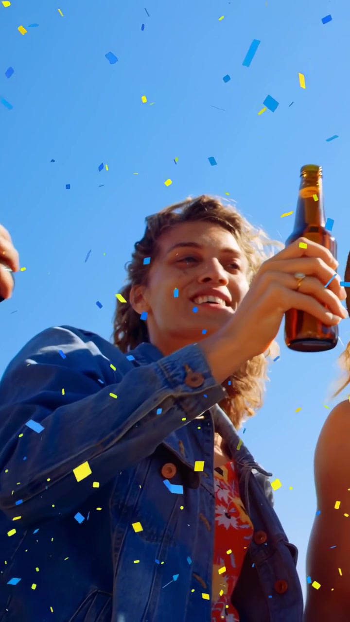 Vertikales Video: Fallendes Konfetti, ein in Jeans gekleideter Erwachsener hebt Bier und trinkt unter blauem Himmel.