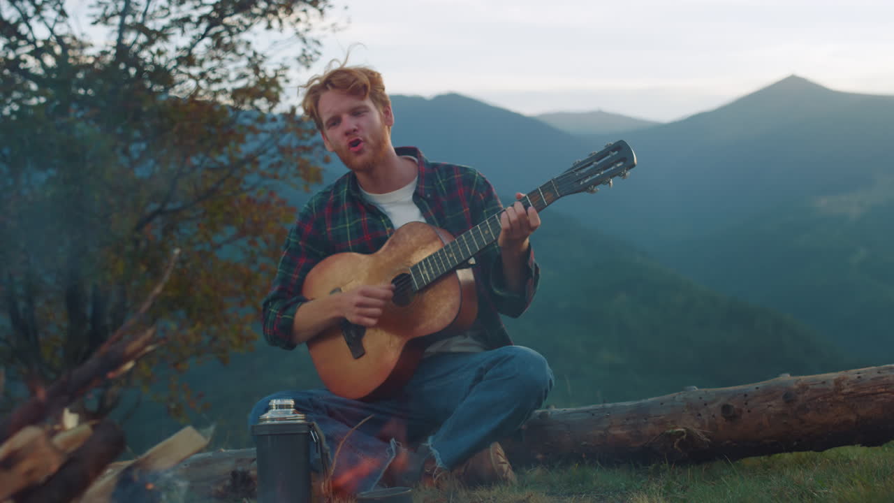 Singender Mann entspannt sich in der Natur im Urlaub. Nahaufnahme: Millennial genießt Lagerfeuer.