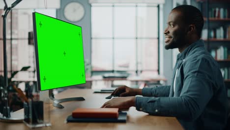 handsome black african american specialist working on desktop computer with green screen mock up display at home living room. freelance man chatting to clients over the internet on social networks.