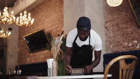 a black person in a black apron and white t-shirt places a reserved sign on a table in a restaurant. table reservation in a restaurant