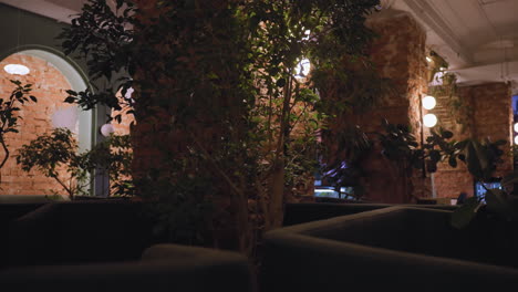 warmly lit room showcases cozy seating area with indoor plants and exposed brick walls. cushions and greenery create serene ambiance while decorative lighting highlights architectural textures and natural accents