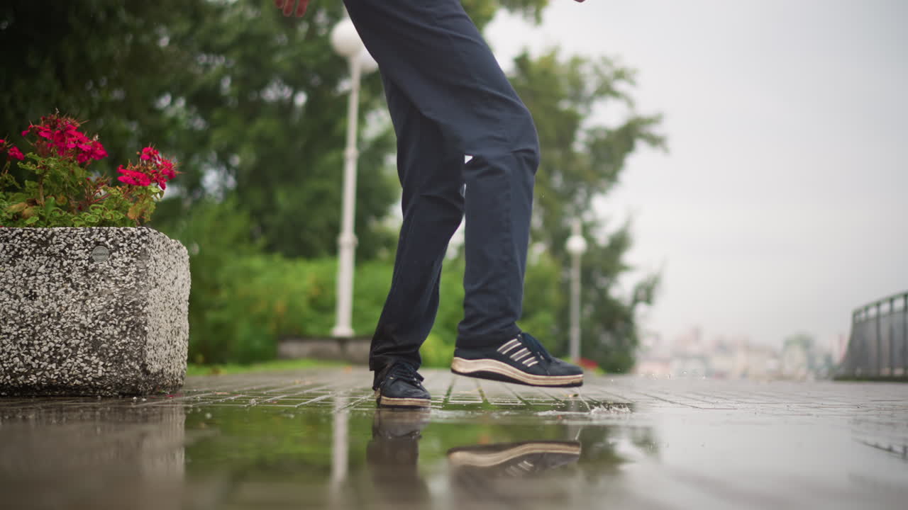 wide step city sidewalk puddle splash, commuter crossing bridge near planter flowers, athletic shoes striking water, visible sole and gait, brisk purposeful pace, overcast sky, cinematic leg motion