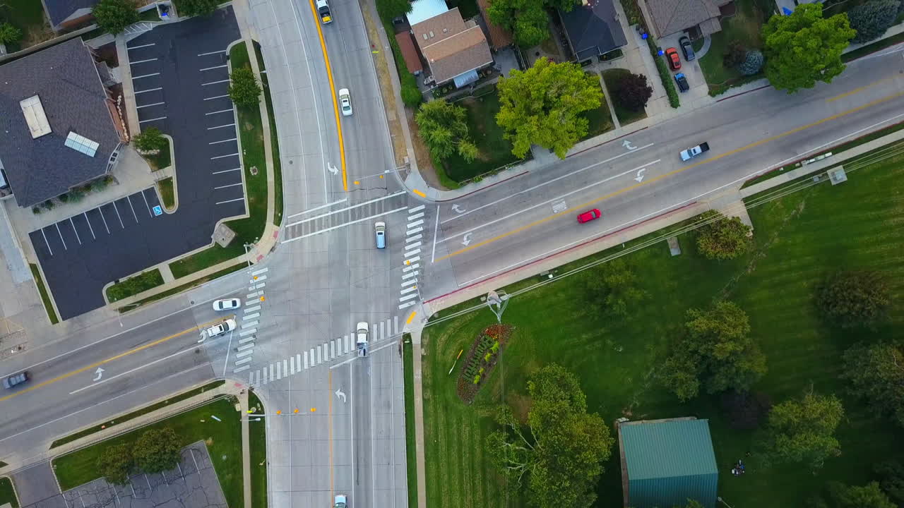 A drone flys over an intersection in a town next to a park as cars drive going their own directions and obeying traffic laws.