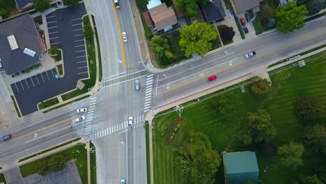 a drone flys over an intersection in a town next to a park as cars drive going their own directions and obeying traffic laws.