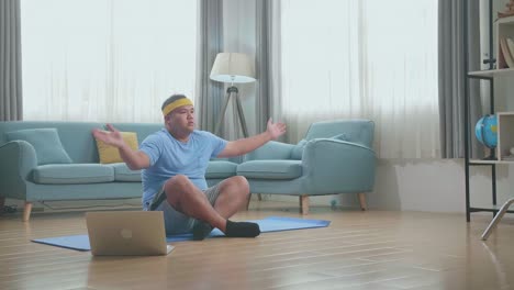 fat asian man on mat looking at laptop and doing yoga in sukhasana pose at home