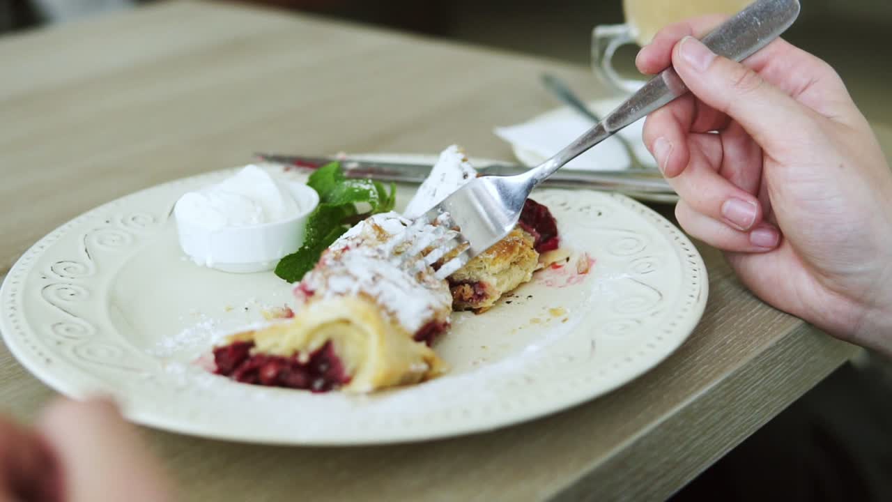 ⁇  티셔츠를 입은 알아보기 힘든 남자가 포크와 칼을 사용하여 레스토랑에서 사막 스트루델을 먹고 있습니다. 슬로우 모션 촬영